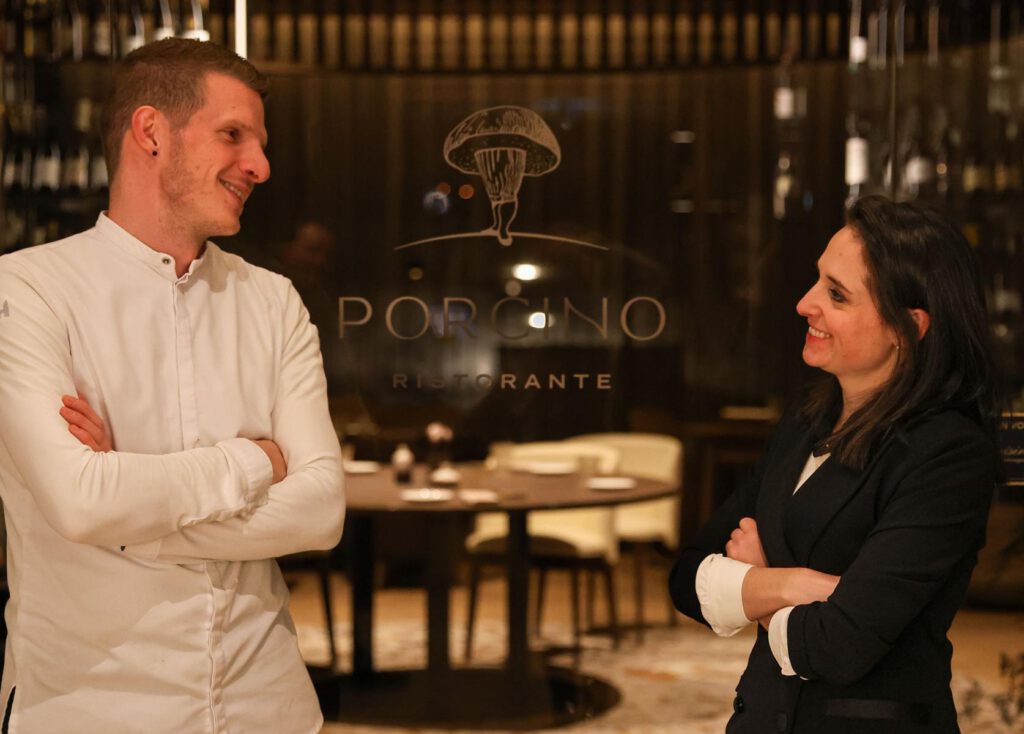 Ein Mann und eine Frau stehen sich lächelnd und mit verschränkten Armen in einem Restaurant gegenüber, an dessen Glastür „Porcino Ristorante“ zu sehen ist.