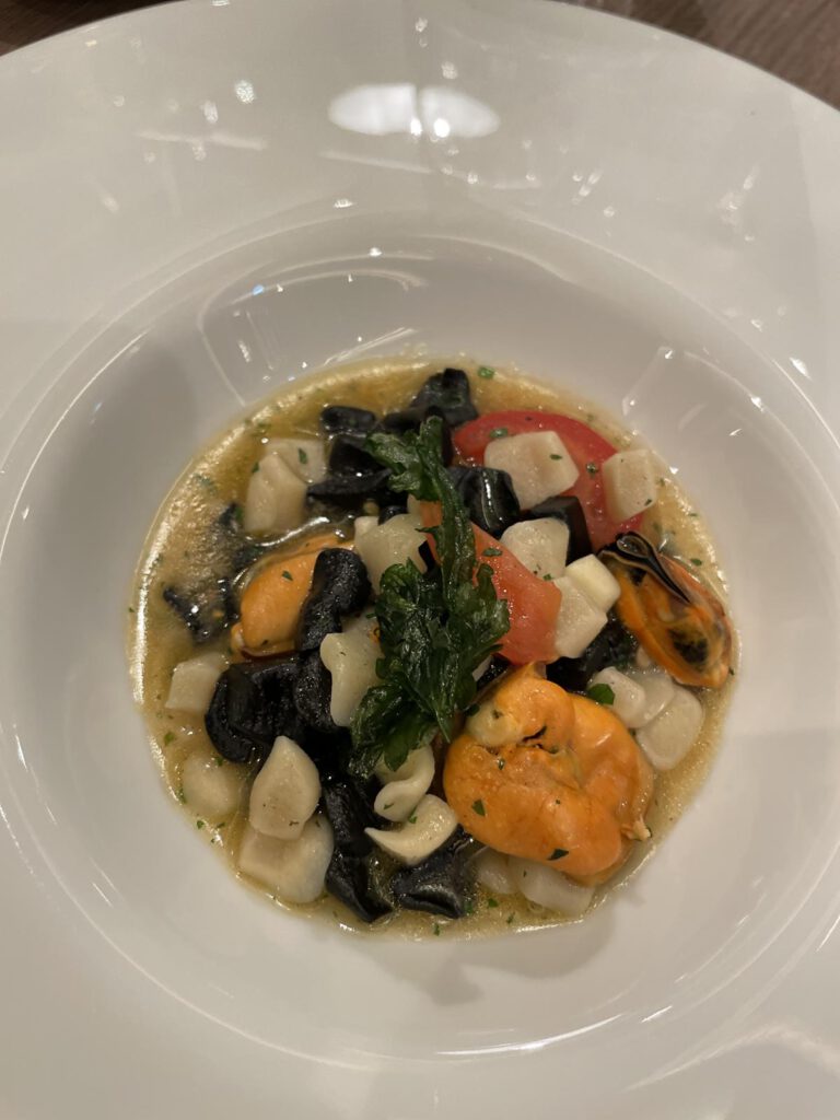 Ein weißer Teller mit einem Meeresfrüchtegericht bestehend aus schwarzen Nudeln, Muscheln, gewürfelten Jakobsmuscheln und Tomaten in einer leichten Brühe, garniert mit einem Zweig grüner Kräuter.