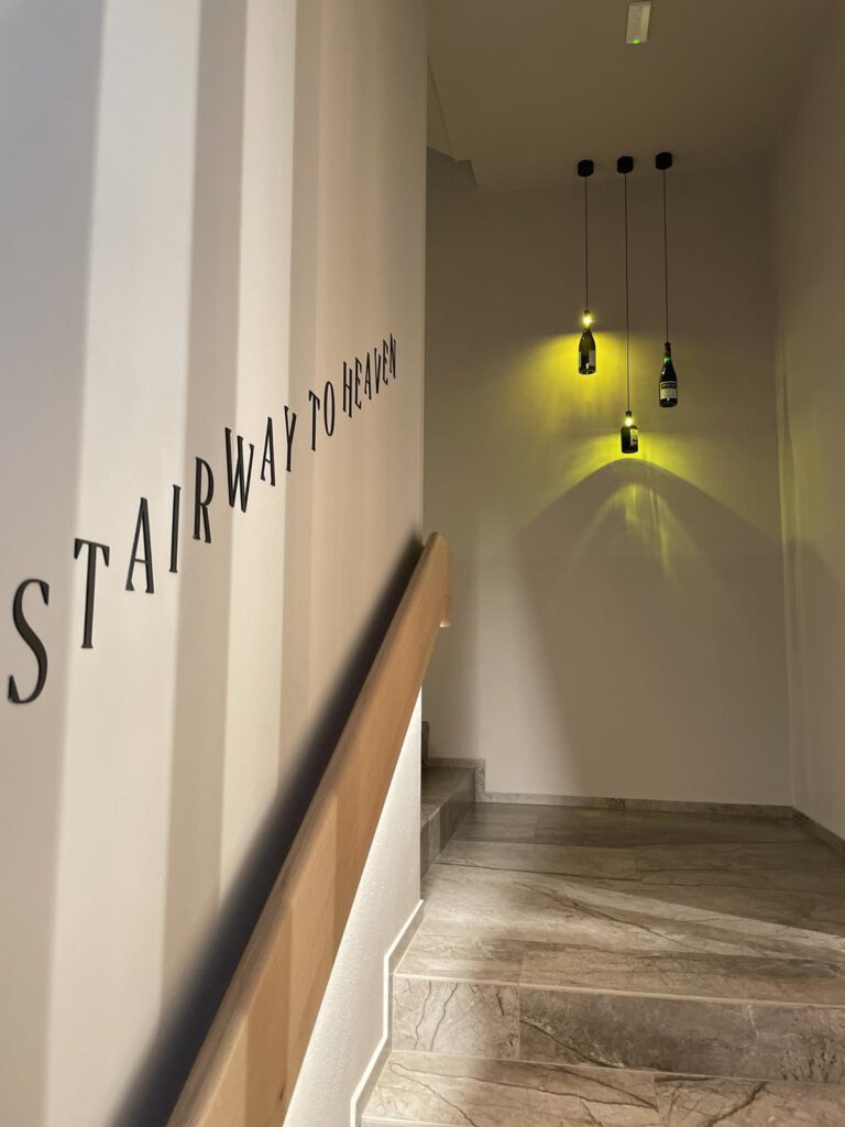 Treppe mit dem Text „Stairway to Heaven“ an der Wand, drei hängende flaschenförmige Lichter beleuchten den Bereich.