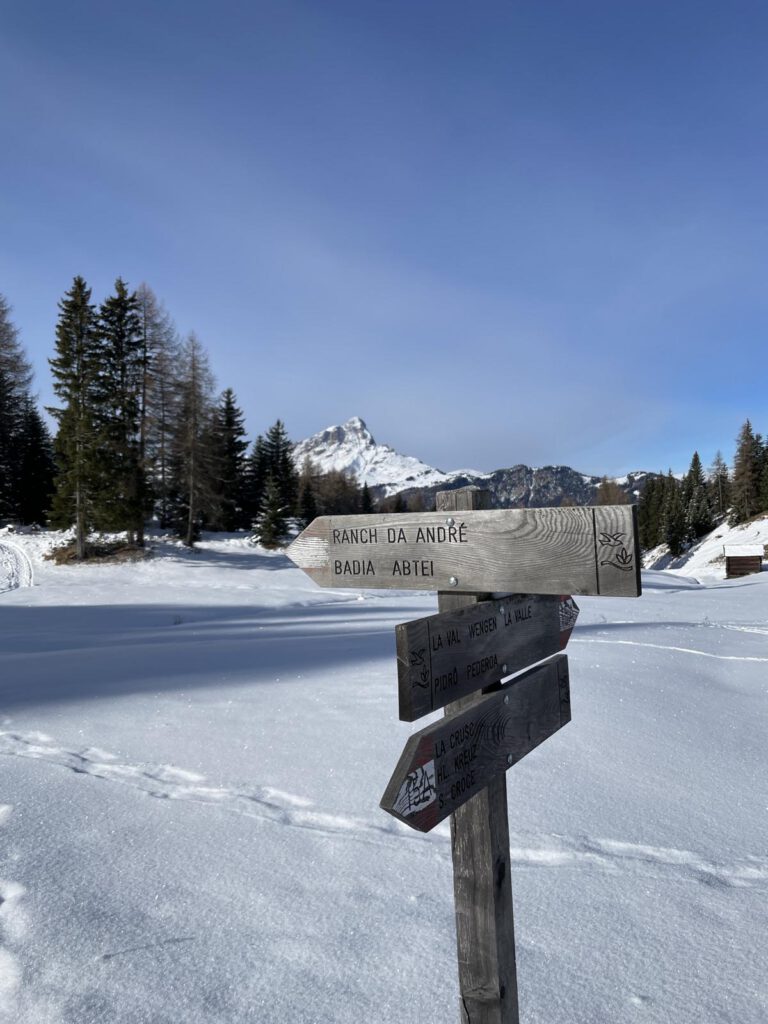 Verschneite Landschaft mit einem hölzernen Wegweiser im Vordergrund, der in verschiedene Richtungen weist. Unter einem klaren blauen Himmel sind schneebedeckte Berge und Bäume zu sehen.