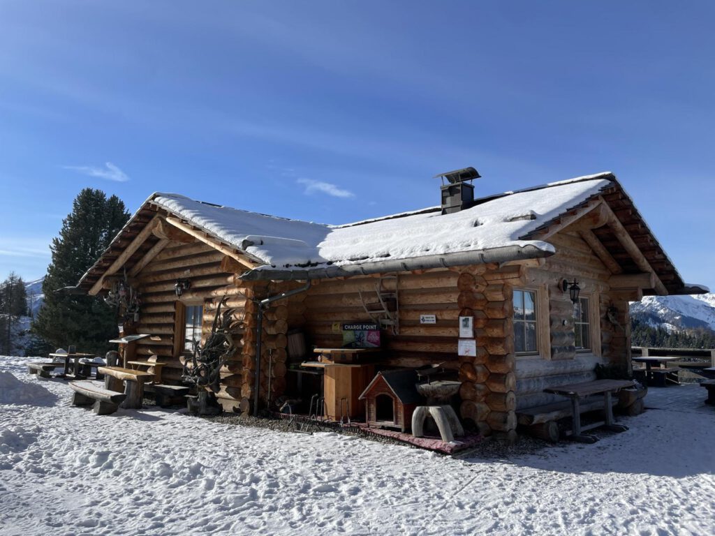 Eine Holzhütte in einer verschneiten Landschaft mit Picknicktischen draußen. Klarer blauer Himmel und umliegende Berge im Hintergrund.