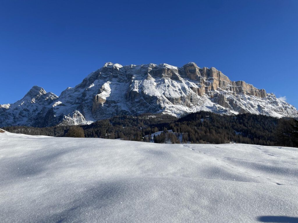 Schneebedeckte Landschaft mit bergiger Kulisse unter klarem blauen Himmel. Der Berg weist felsige Strukturen auf, im Vordergrund ein glattes, schneebedecktes Feld. Vereinzelte Bäume sind sichtbar.