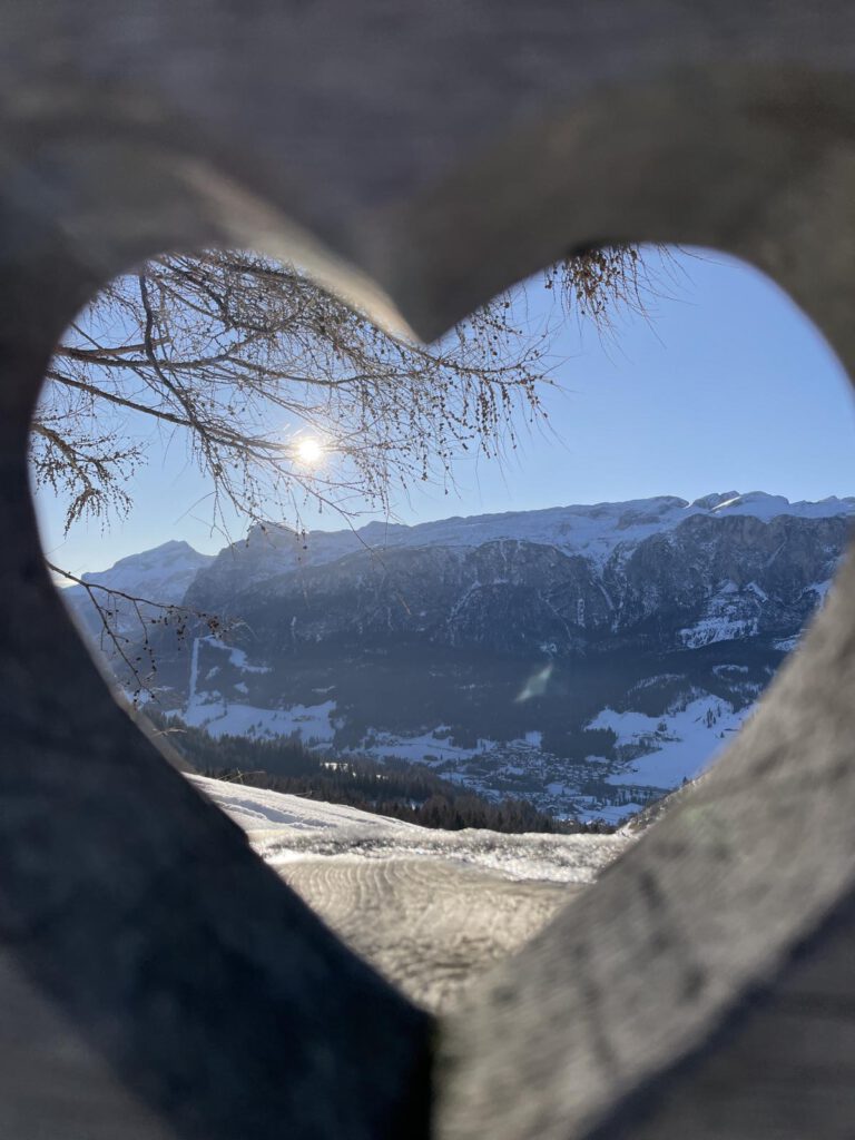 Schneebedeckte Berge und eine strahlende Sonne, betrachtet durch einen herzförmigen Holzrahmen. Äste sind sichtbar und im Hintergrund ist ein klarer, blauer Himmel zu sehen.
