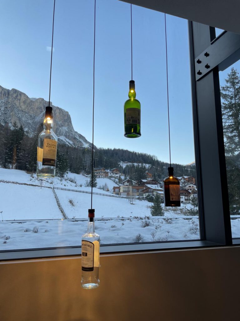 Vier Glasflaschen hängen kopfüber als Pendelleuchten vor einer verschneiten Berglandschaft, die durch ein großes Fenster sichtbar ist.