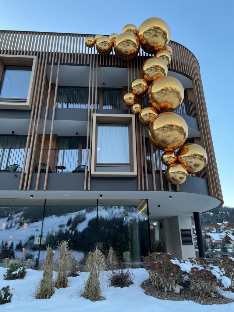 Ein modernes Gebäude mit großen, reflektierenden Goldkugeln an der Fassade. Schnee bedeckt den Boden und die Landschaft im Hintergrund.