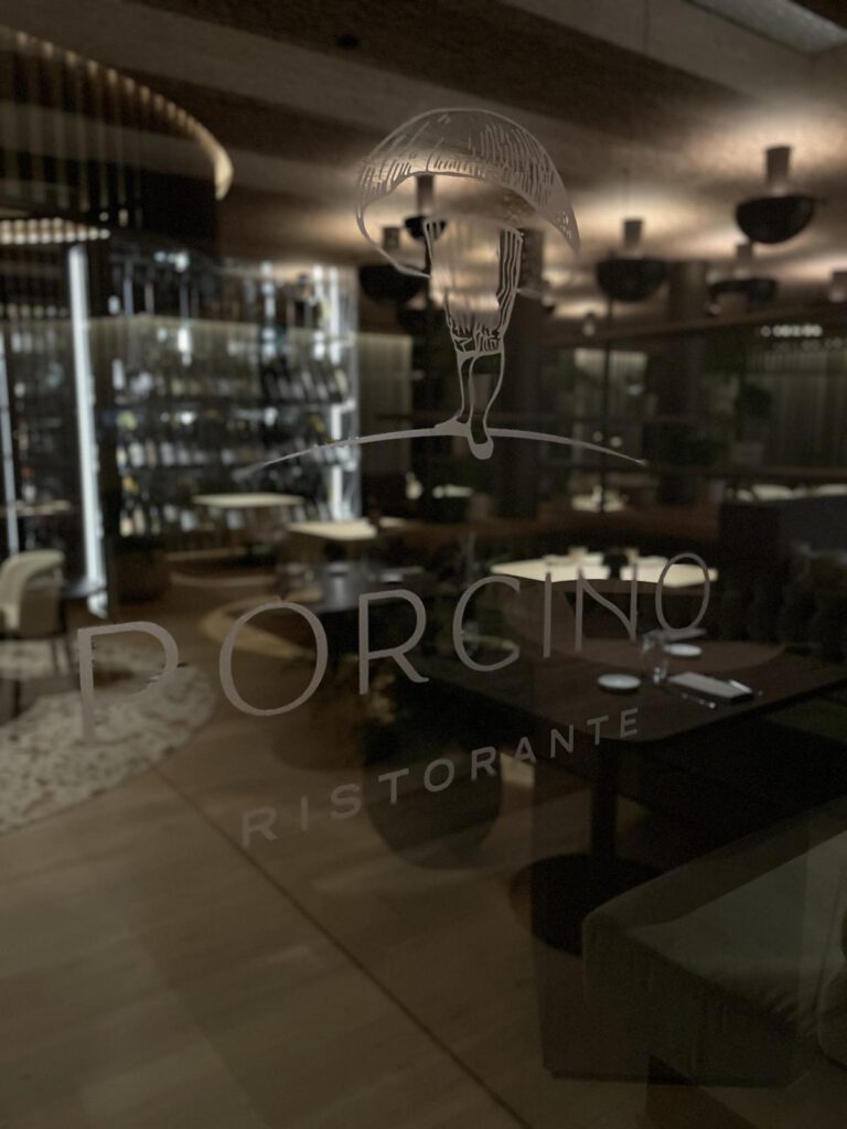 Schwach beleuchteter Restaurantinnenraum mit Fokus auf der Glastür mit dem eingravierten Namen „Porcino Ristorante“. Im Hintergrund sind Tische, Stühle und Weinregale zu sehen.