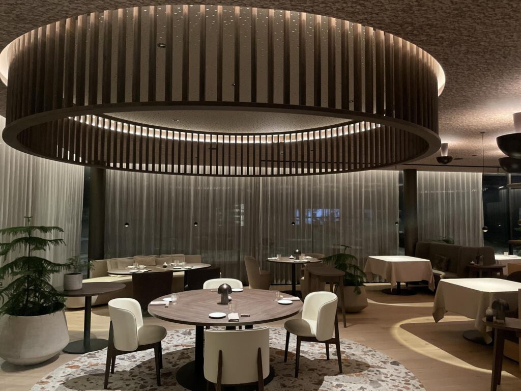 Modernes Restaurant-Interieur mit runden Tischen, weißen Stühlen und einer großen runden Deckenleuchte. Sanftes Ambientelicht und raumhohe Vorhänge schaffen eine elegante Atmosphäre.