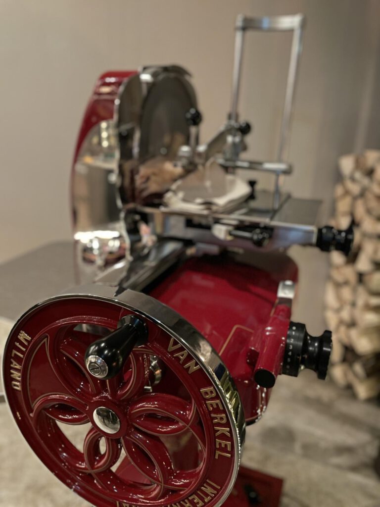 Rote Vintage-Schneidemaschine mit „Van Berkel“ auf dem Rad, auf einem Tisch platziert. Im Hintergrund ist gestapeltes Brennholz zu sehen.