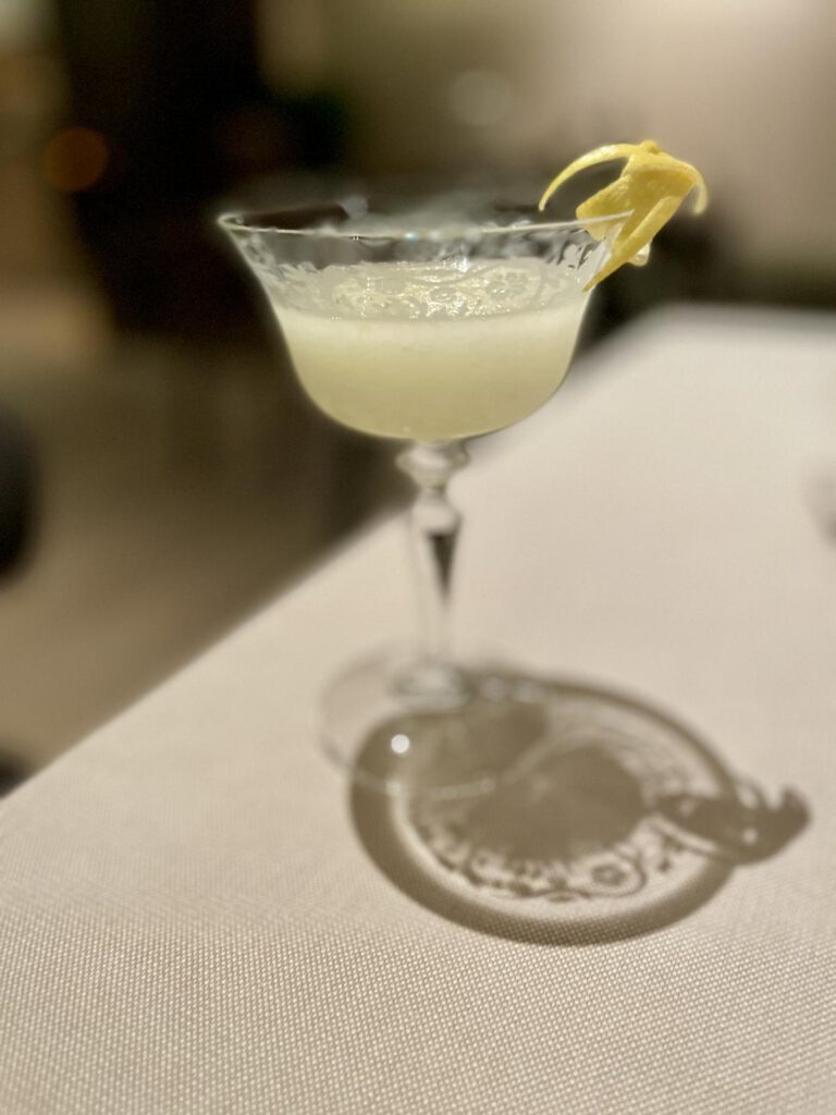 Ein Cocktail in einem Glas mit Zuckerrand und Zitronenspirale als Garnitur steht auf einer Tischdecke.