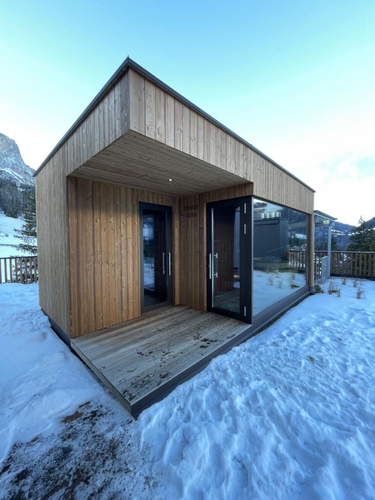Eine kleine, moderne Holzhütte mit großen Glasfenstern, gelegen in einer verschneiten Landschaft. Das Gebäude verfügt über einen überdachten Holzeingang.