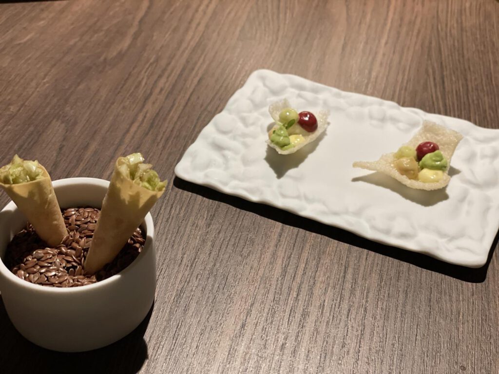 Zwei knusprige Waffeln mit grüner Füllung in einer Tasse mit Körnern neben einem Teller mit zwei knusprigen Häppchen, garniert mit kleinen bunten Garnituren auf einem Holztisch.