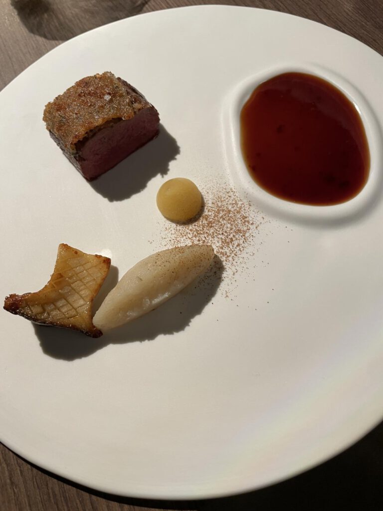 Gourmetgericht mit einer Scheibe Fleisch, Soße, weißer Mousse, kleinem runden Gel und einem Stück gegrilltem Pilz auf einem weißen Teller.
