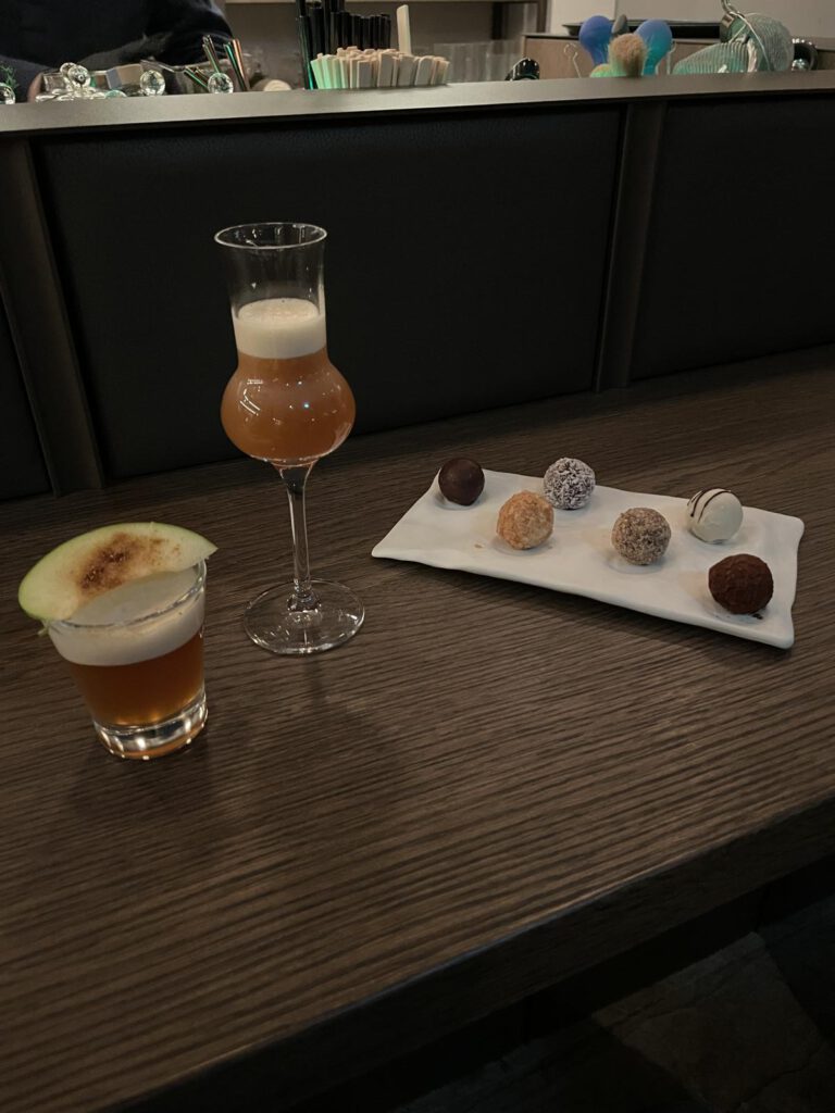 Zwei Cocktails auf einem Tisch, garniert mit einer Apfelscheibe. Daneben steht ein rechteckiger Teller mit sechs verschiedenen Trüffeln.