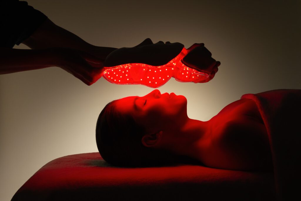 Eine Person liegt unter einer roten LED-Lichttherapiemaske, der Raum ist schwach beleuchtet.