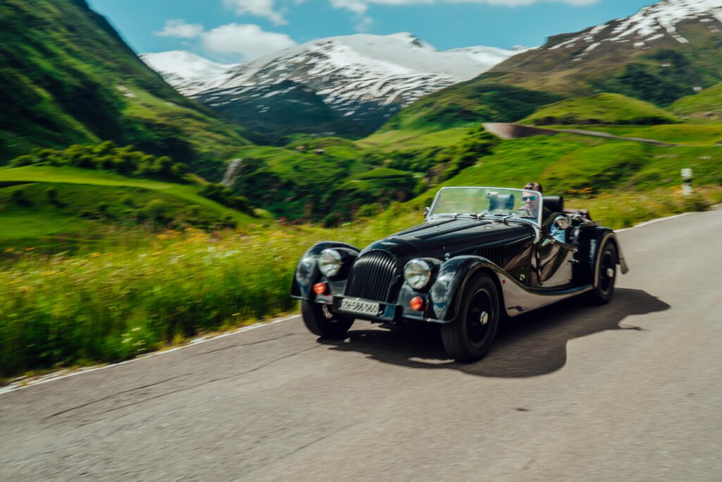 Ein schwarzes Oldtimer-Cabrio fährt unter einem klaren Himmel auf einer malerischen Bergstraße mit grünen Hügeln und schneebedeckten Gipfeln im Hintergrund.