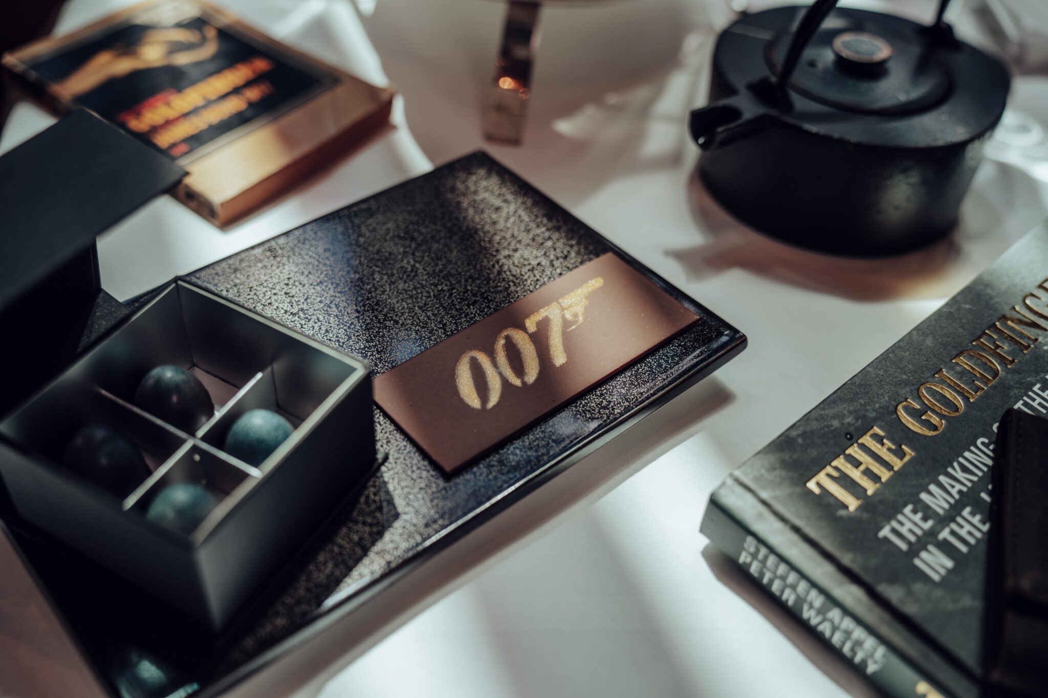 Eine Nahaufnahme eines Tisches mit einem „007“-Schild, einem Buch mit dem Titel „Akte Goldfinger“, einer Teekanne und einer Schachtel mit vier dunklen Kugeln.