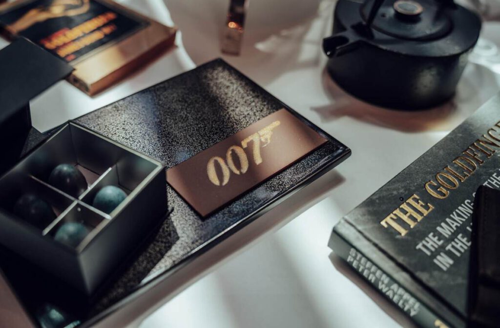 Eine Nahaufnahme eines Tisches mit einem „007“-Schild, einem Buch mit dem Titel „Akte Goldfinger“, einer Teekanne und einer Schachtel mit vier dunklen Kugeln.