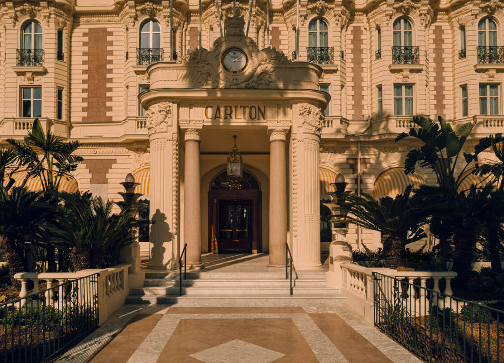 Der Eingang des Carlton Hotels besticht durch verzierte Säulen, eine große Uhr über dem Eingang und umgebende Palmen im warmen Sonnenlicht.