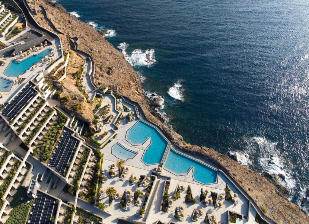 Luftaufnahme eines modernen Badeortes mit mehreren Pools, Reihenhäusern und Solarmodulen neben einer felsigen Küste und dem Meer.