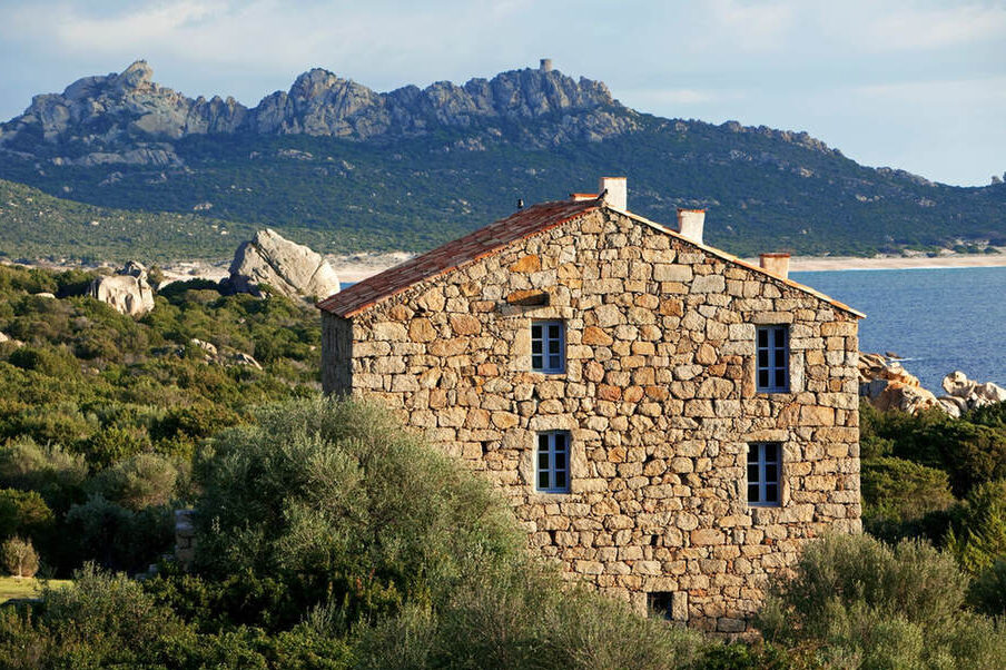 Ein Steinhaus steht inmitten grüner Sträucher mit Blick auf das Meer, im Hintergrund felsige Hügel und ferne Berge unter einem blauen Himmel.