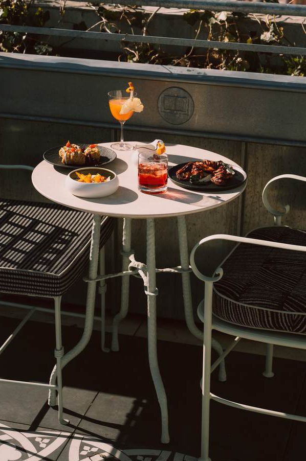 Ein runder weißer Tisch mit zwei Stühlen bietet Platz für zwei Cocktails, kleine Schalen mit Vorspeisen und einen Teller mit Gegrilltem, der im Freien im Sonnenlicht steht.