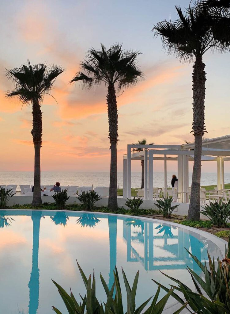 Eine heitere Poolszene bei Sonnenuntergang mit Palmen, einem ruhigen Pool, in dem sich der Himmel spiegelt, und zwei Personen in der Nähe von weißen Außenstrukturen am Meer.