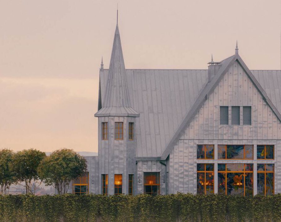 Ein metallisches, turmartiges Haus mit hohen Fenstern und spitzem Dach steht in der Abenddämmerung hinter einer Reihe ordentlich gestutzter Bäume und Hecken.