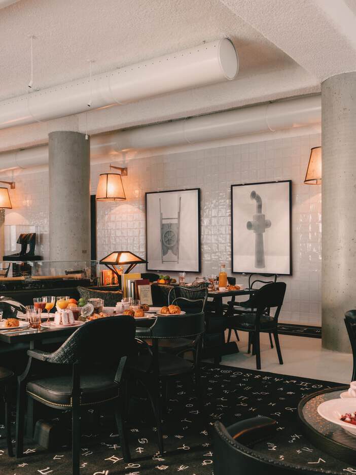 Modernes Restaurantinterieur mit gedeckten Esstischen, schwarzen Stühlen, gerahmter abstrakter Kunst an einer weiß gefliesten Wand und Umgebungsbeleuchtung.