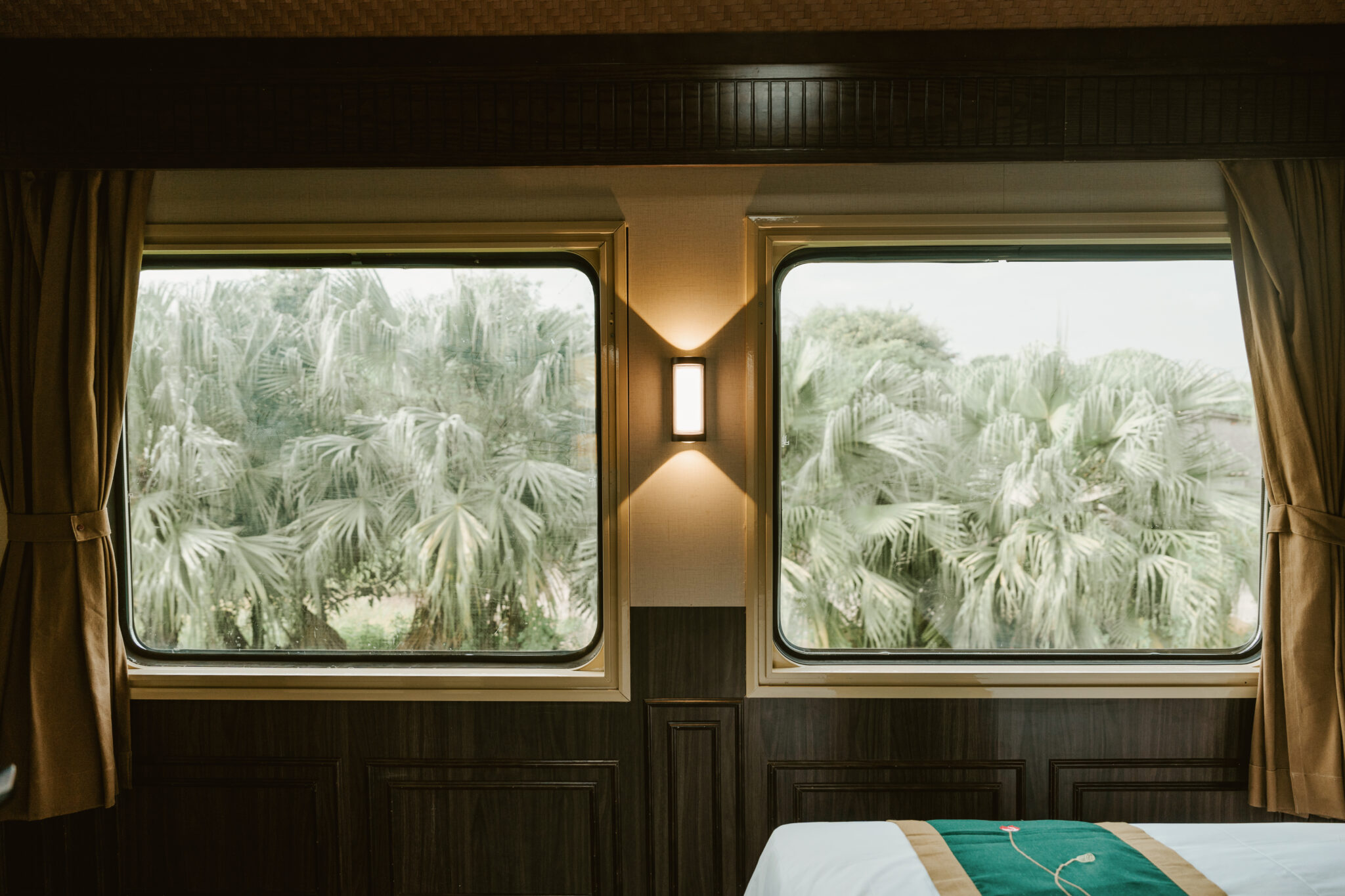 Zwei große Fenster in einem Zugabteil geben den Blick auf Palmen frei, dazwischen eine Wandlampe und im Vordergrund ein Teil eines Bettes.