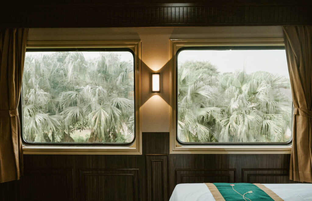 Zwei große Fenster in einem Zugabteil geben den Blick auf Palmen frei, dazwischen eine Wandlampe und im Vordergrund ein Teil eines Bettes.
