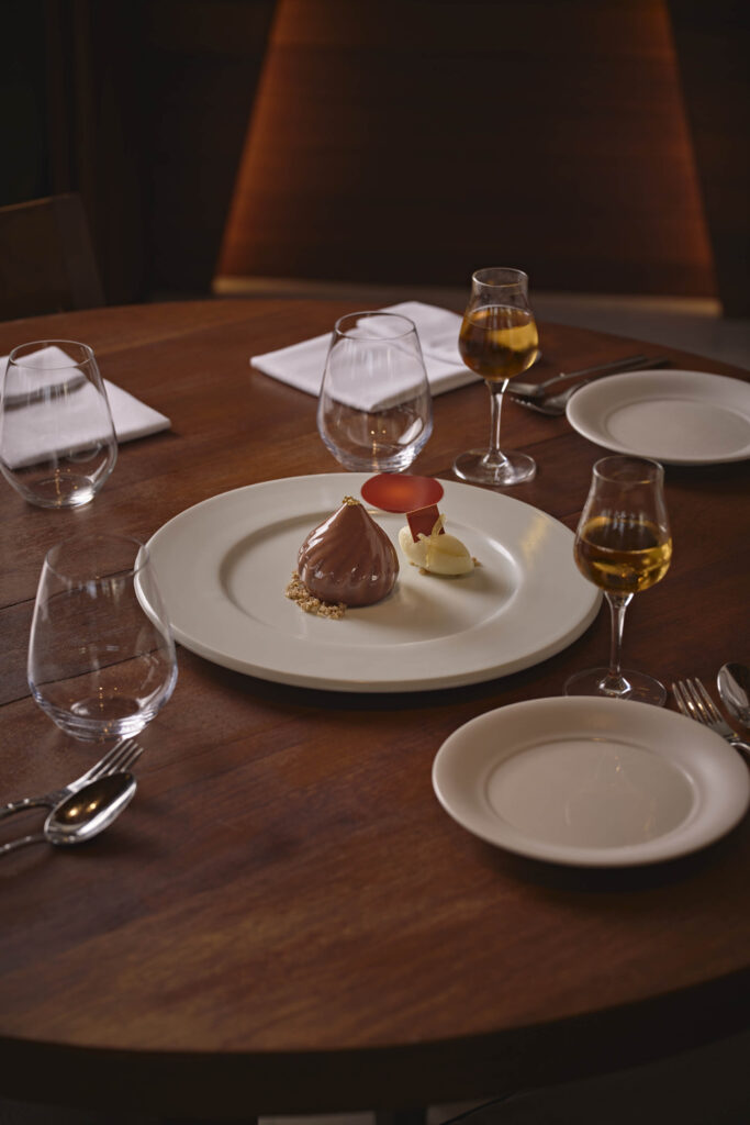 Ein edel angerichtete Nachspeise auf einem weißen Teller, umringt von leeren Wassergläsern und zwei gefüllten Weingläsern.