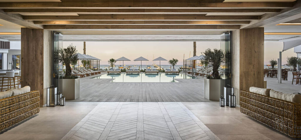 Blick aus einer modernen Hotellobby auf einen Poolbereich mit Liegestühlen und Sonnenschirmen, im Hintergrund ist das Meer zu sehen.