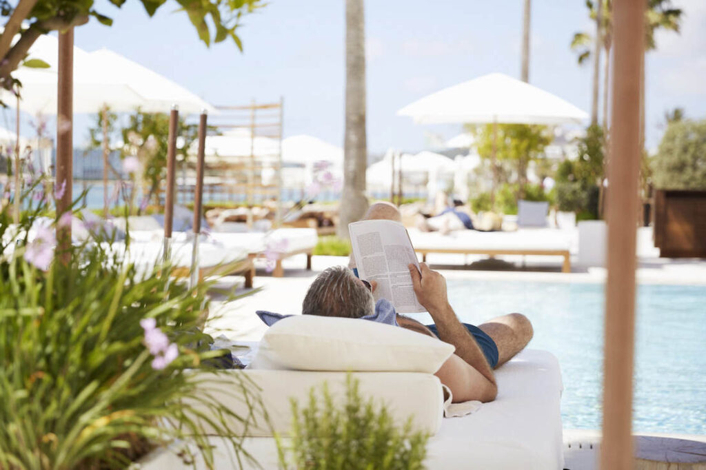 Eine Person liegt auf einem Liegestuhl an einem Pool und liest ein Buch. Im Vordergrund befinden sich Pflanzen, im Hintergrund weiße Sonnenschirme und weitere Liegestühle.