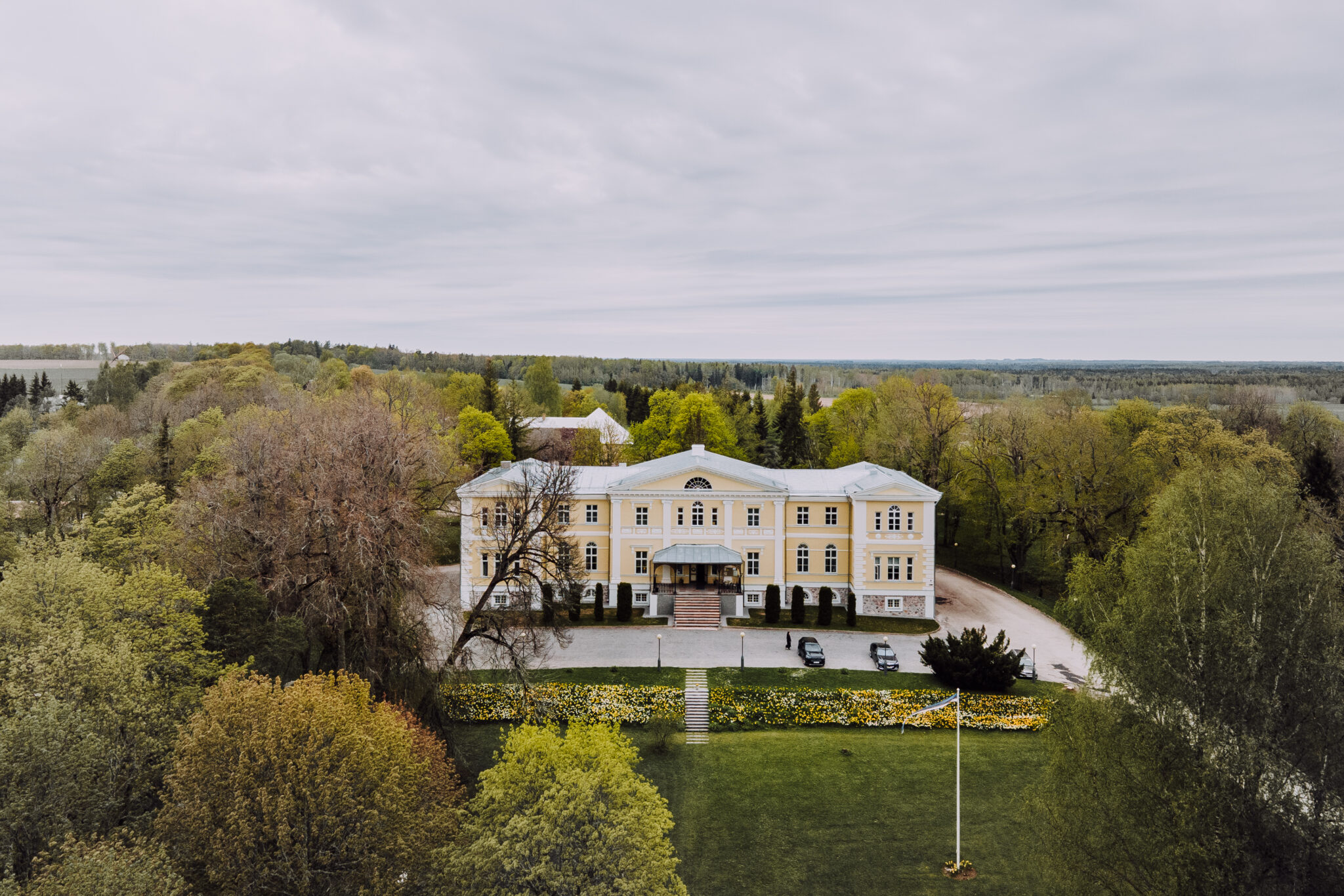 Ein großes, hellgelbes Gebäude mit einer symmetrischen Fassade ist von Bäumen und Gärten unter einem bedeckten Himmel umgeben. Drei Autos sind vor dem Eingang geparkt.