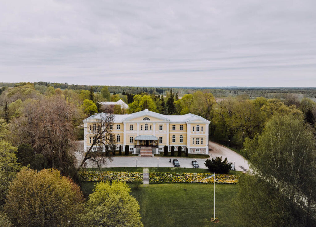 Ein großes, hellgelbes Gebäude mit einer symmetrischen Fassade ist von Bäumen und Gärten unter einem bedeckten Himmel umgeben. Drei Autos sind vor dem Eingang geparkt.