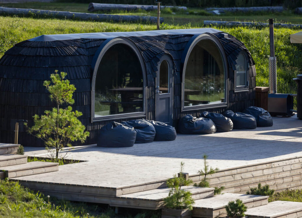 Eine kleine, geschwungene Hütte mit großen Bogenfenstern, schwarzen Schindeln und mehreren schwarzen Sitzsäcken auf einer von Grün umgebenen Holzterrasse.