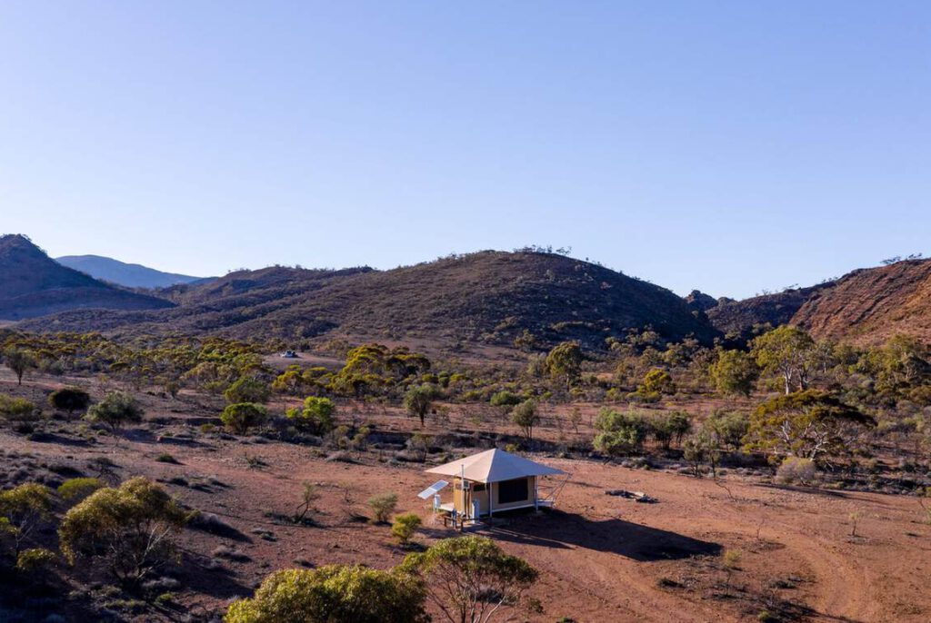 Eine kleine Zelthütte steht allein in einer trockenen, offenen Landschaft mit vereinzelten Bäumen und entfernten Hügeln unter einem klaren blauen Himmel.