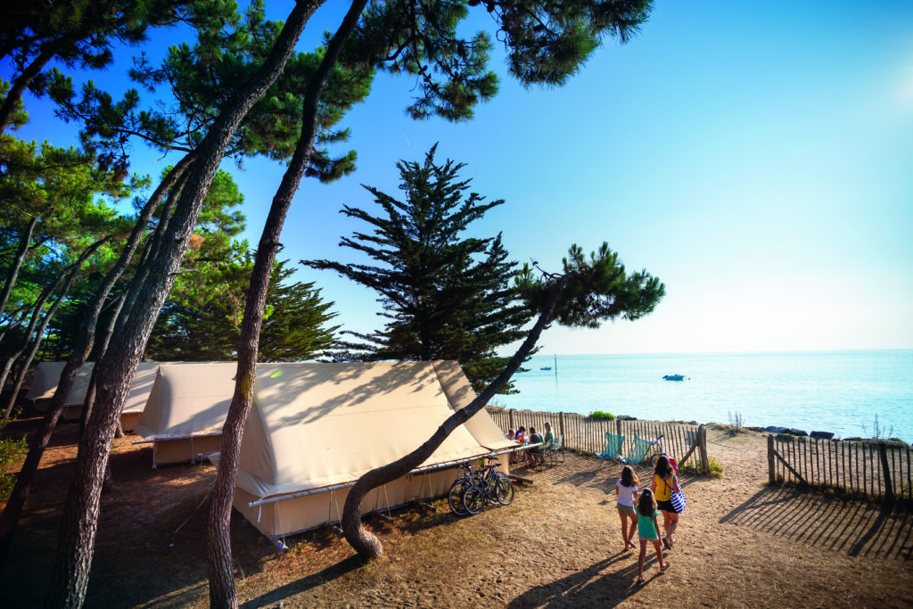 Drei Personen spazieren in der Nähe von großen Zelten und Fahrrädern zwischen Bäumen auf einer Sandfläche am Meer, mit klarem Himmel und ruhigem Wasser im Hintergrund.