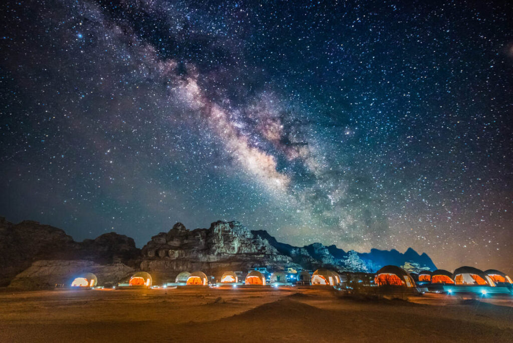 Eine Reihe von beleuchteten Wüstenzelten steht unter einer felsigen Bergkette mit der Milchstraße und einem sternenübersäten Nachthimmel über sich.