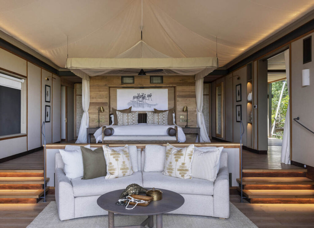 Ein modernes, gehobenes Schlafzimmer mit Himmelbett, neutralem Sofa, dekorativen Kissen, Holzakzenten und sanfter Beleuchtung auf den Stufen, die zum Bettbereich führen.