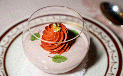 Eine gläserne Dessertschale mit rosafarbener Mousse, gekrönt von einem Strudel aus Schokoladencreme, Blattgold und zwei kleinen grünen Blättern, auf einem dekorativen Teller.