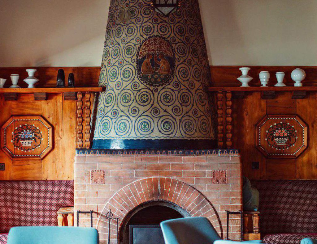 In der gemütlichen Lounge gibt es einen großen, verzierten, konischen Kamin, blaue Sessel, Holzvertäfelung und weiße Keramik auf einem Kaminsims.