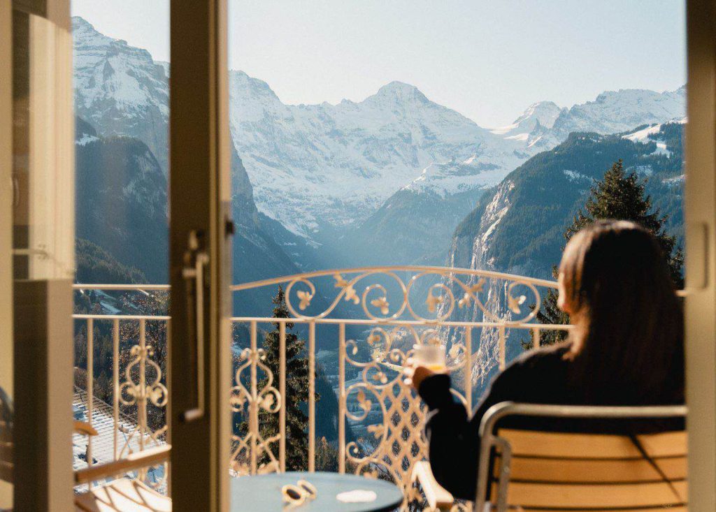 Eine Person sitzt mit einem Getränk in der Hand auf einem Balkon und blickt auf eine malerische Bergkette mit schneebedeckten Gipfeln und einem klaren Himmel.