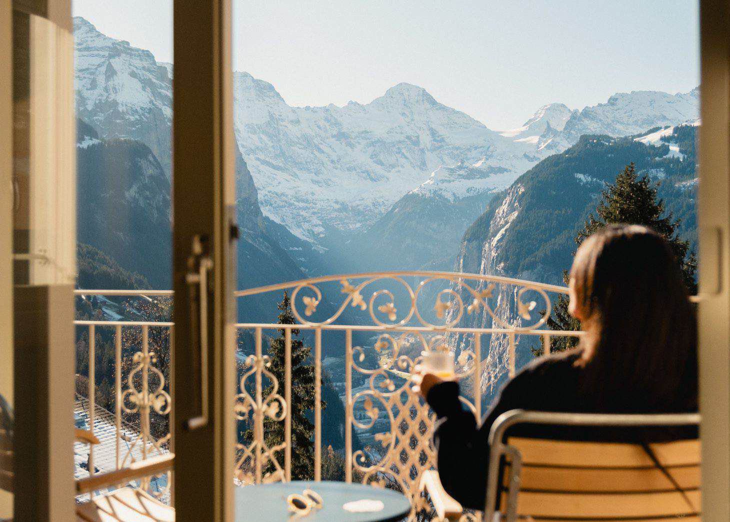 Eine Person sitzt mit einem Getränk in der Hand auf einem Balkon und blickt auf eine malerische Bergkette mit schneebedeckten Gipfeln und einem klaren Himmel.