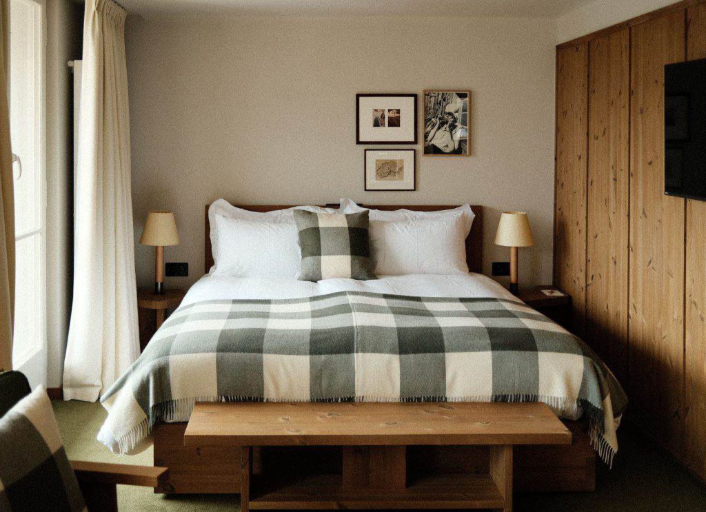 Ein ordentlich gemachtes Bett mit einer karierten Decke und einem passenden Kissen, eingerahmt von zwei Nachttischen mit Lampen, Holzmöbeln, Wandkunst über dem Bett und einem Flachbildfernseher an der rechten Wand.