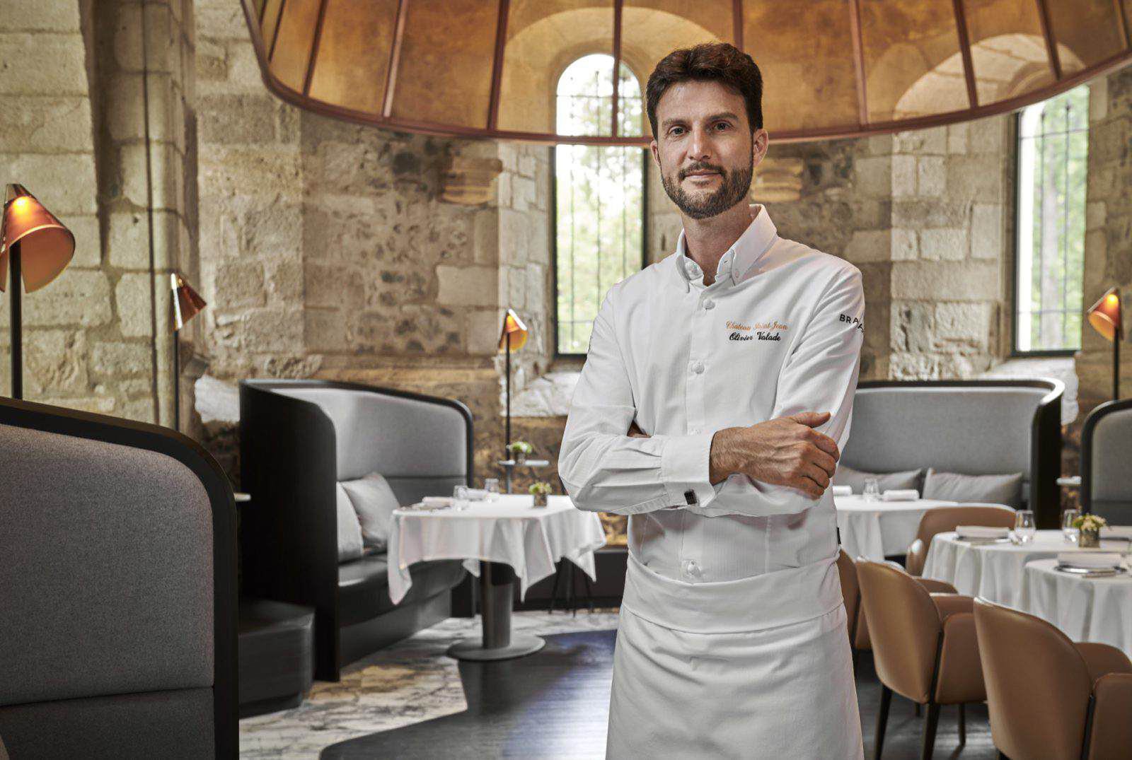 Ein Koch in weißer Uniform steht mit verschränkten Armen in einem eleganten, steingemauerten Restaurant mit gedeckten Tischen und moderner Bestuhlung.