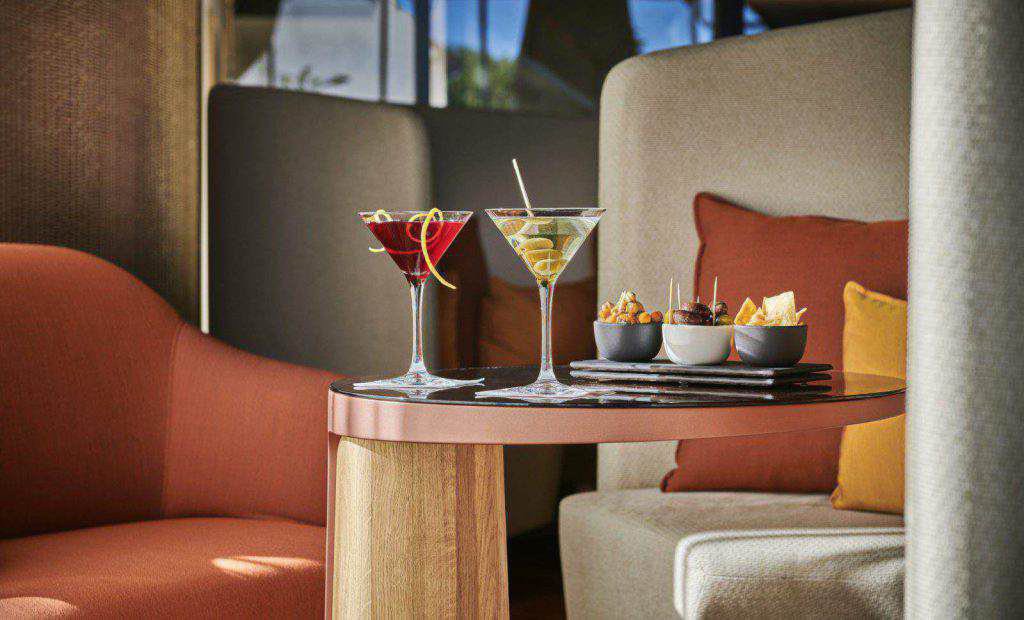 Zwei Cocktails und verschiedene Snacks sind auf einem runden Holztisch zwischen orangefarbenen und beigen Sitzkissen in einer Lounge angeordnet.