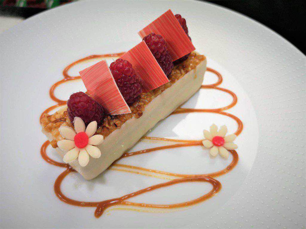 Ein rechteckiges Dessert mit Himbeeren und rosa Schokoladenstückchen, garniert mit Karamellsauce und zwei weißen Schokoladenblumen auf einem weißen Teller.