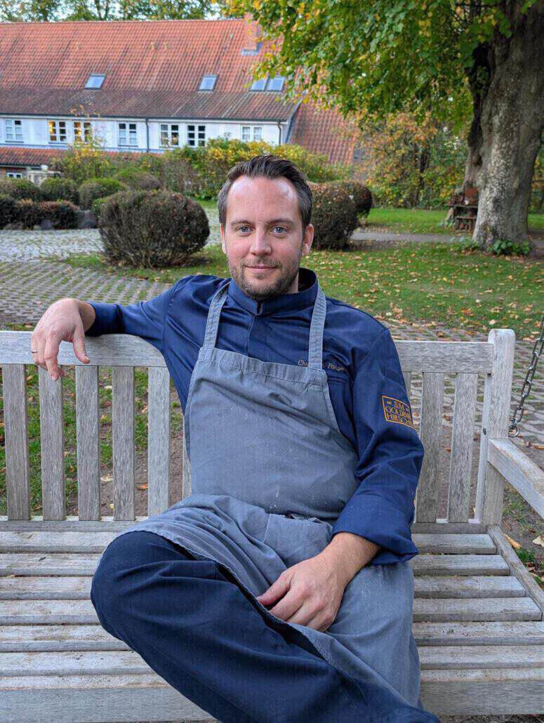 Ein Koch in blauer Arbeitsschürze und blauem Kochhemd sitzt auf einer grauen Holzbank. Im Hintergrund sind ein altes Gutshaus sowie verschiedene Bäume und Sträucher im Herbst zu sehen.