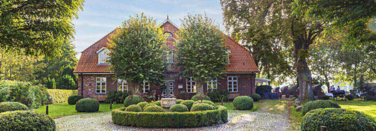 Blick auf ein altes Gutshaus mit rotem Dach und roten Backsteinen im Herbst. Davor sind zwei Bäumen angeordnet, die Sonne scheint von rechts oben durch eine alte Baumkrone und ein runder, bepflanzter Platz ist im Vordergrund zu sehen.